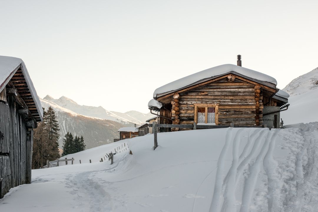 Snow-Covered Log Cabin Nesting on Alpine Slope with Soft Morning Light and Mountain Peaks