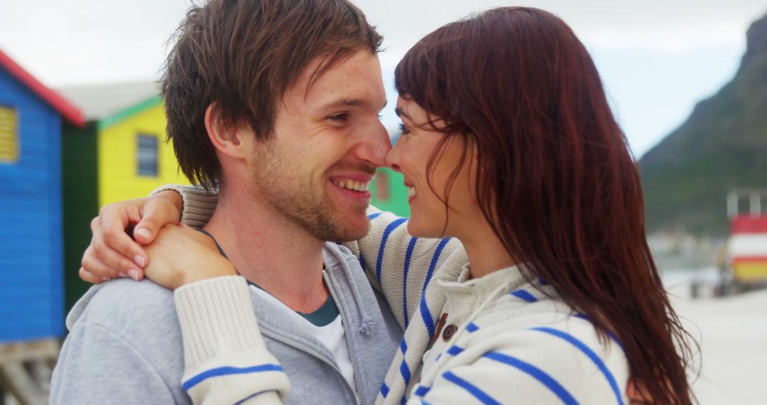 A couple enjoys a romantic embrace on a picturesque beach with vibrant huts. - Free Images, Stock Photos and Pictures on Pikwizard.com