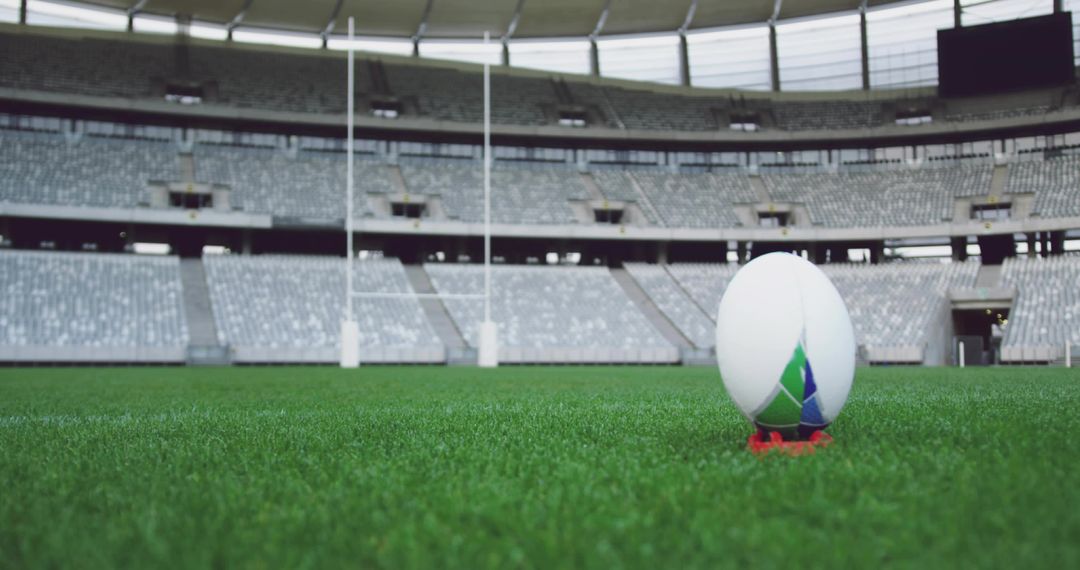 Rugby ball resting on kicking tee in empty stadium with goalposts and scoreboard