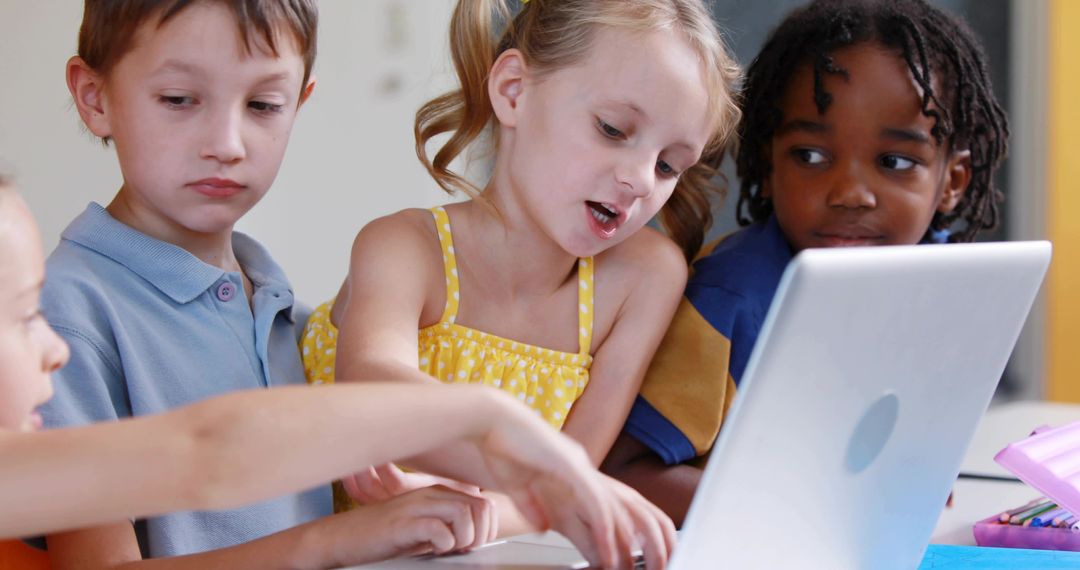 Children collaborating around laptop in elementary classroom, blonde girl pointing