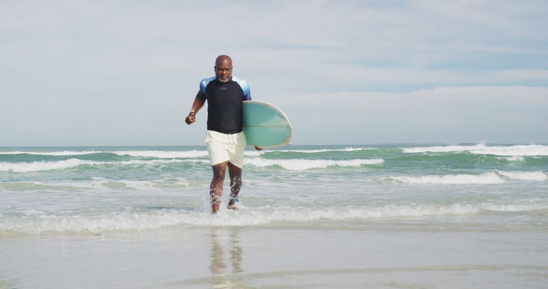 African american senior man walking on a beach holding surfboard and running out of the sea - Free Images, Stock Photos and Pictures on Pikwizard.com
