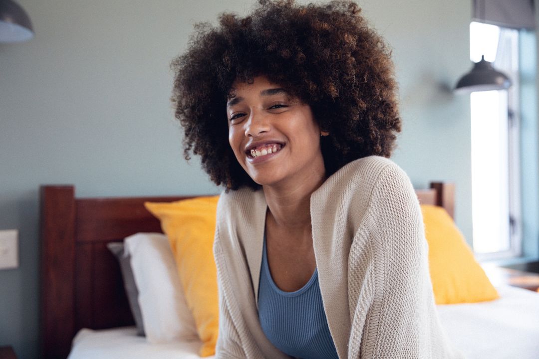 Portrait of smiling biracial young woman with afro hair sitting on bed against wall, copy space - Free Images, Stock Photos and Pictures on Pikwizard.com