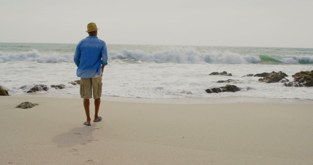 Man in Casual Attire Strolls on Serene Beachfront with Waves