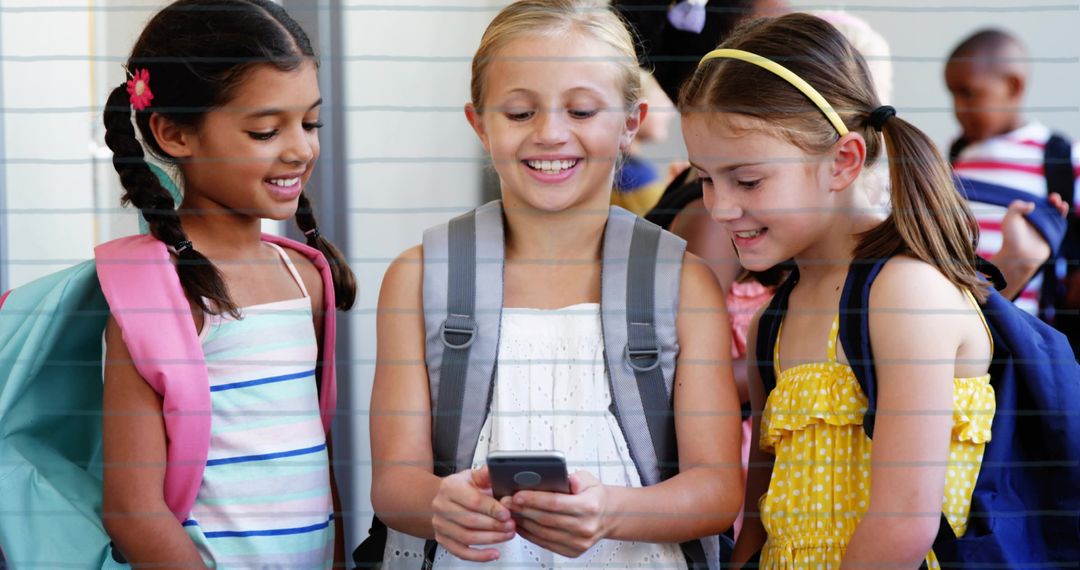 Smiling schoolgirls gathering around smartphone in school corridor with backpacks