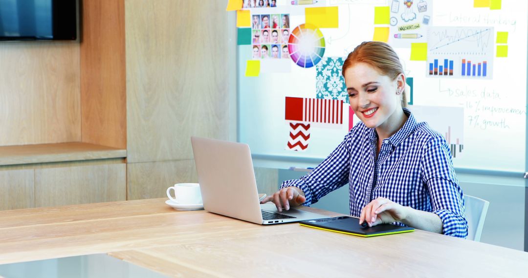 Smiling Woman Using Laptop at Modern Office Desk with Colorful Charts on Wall - Free Images, Stock Photos and Pictures on Pikwizard.com