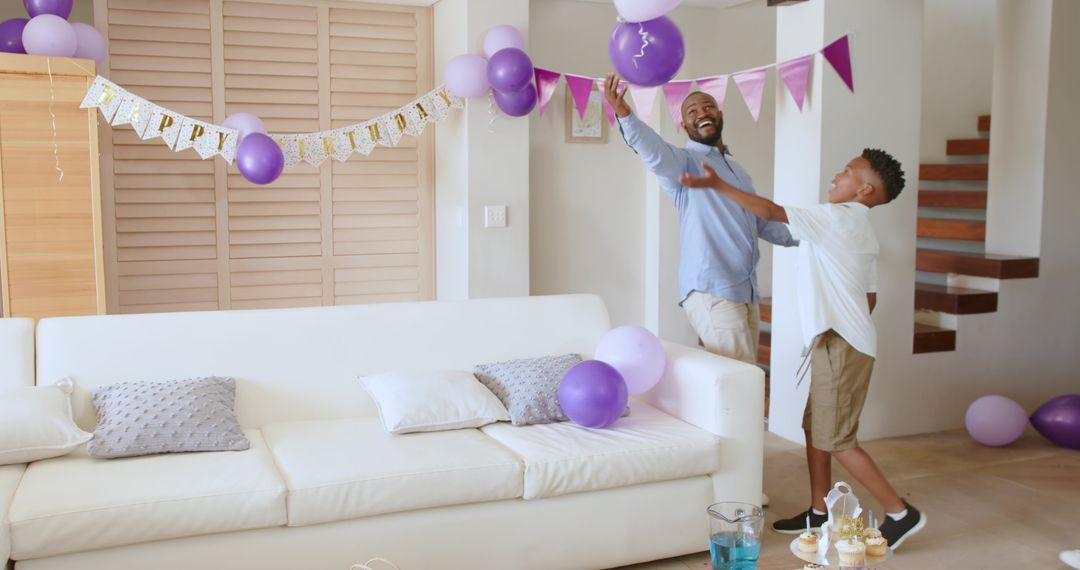 African American Dad and Son Celebrating Birthday Reaching for Purple Balloons in Cozy Living Room