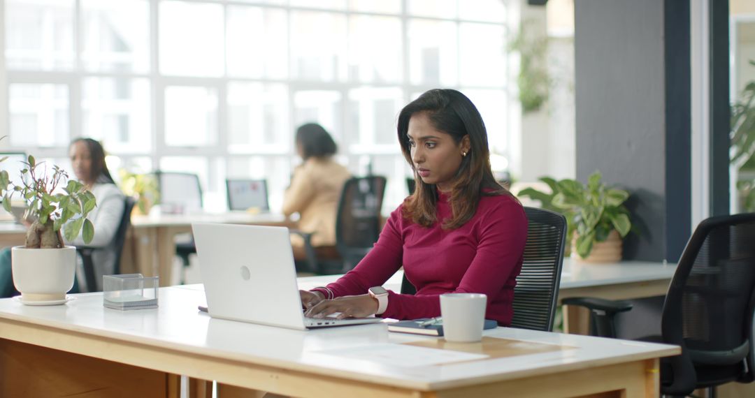 Indian woman working on laptop in modern open-plan office with plants and colleagues