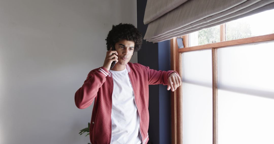 Young man talking on smartphone leaning on window sill wearing red bomber jacket