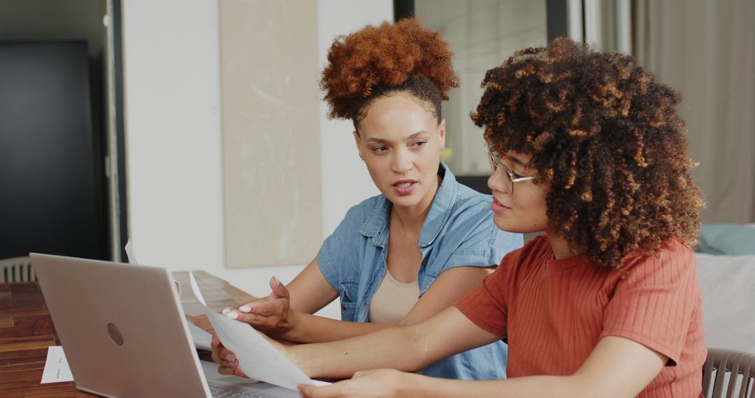 Two women collaborating over laptop and documents, focused in cozy home workspace