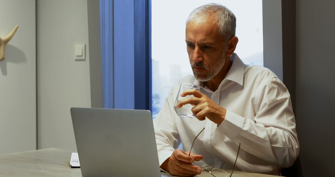 Focused Senior Man Working on Laptop and Drinking Water in Office - Free Images, Stock Photos and Pictures on Pikwizard.com