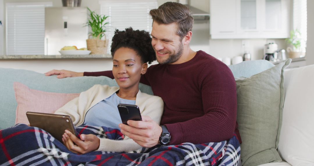 Image of happy diverse couple embracing and using electronic devices on sofa - Free Images, Stock Photos and Pictures on Pikwizard.com