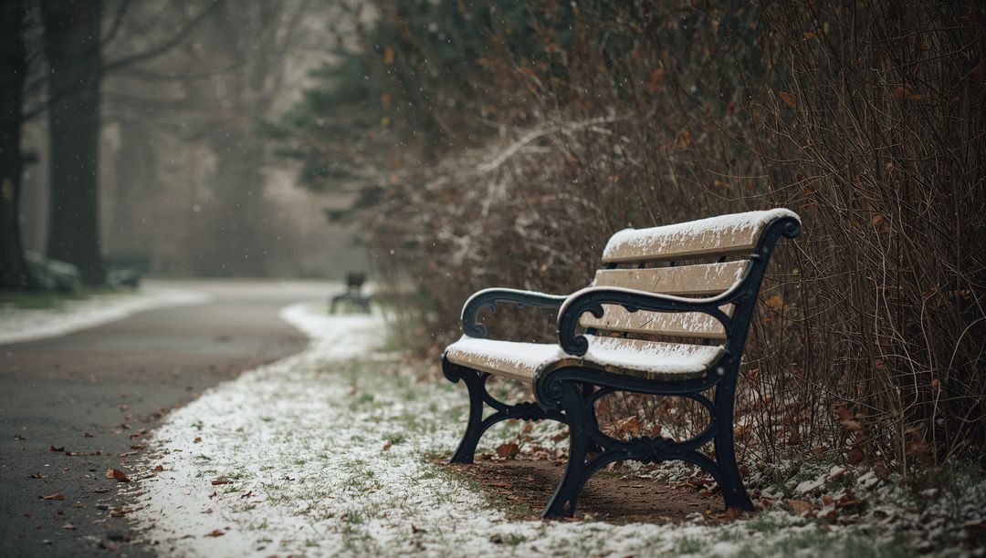 Wooden bench collecting light snow on winding park path in quiet foggy winter morning