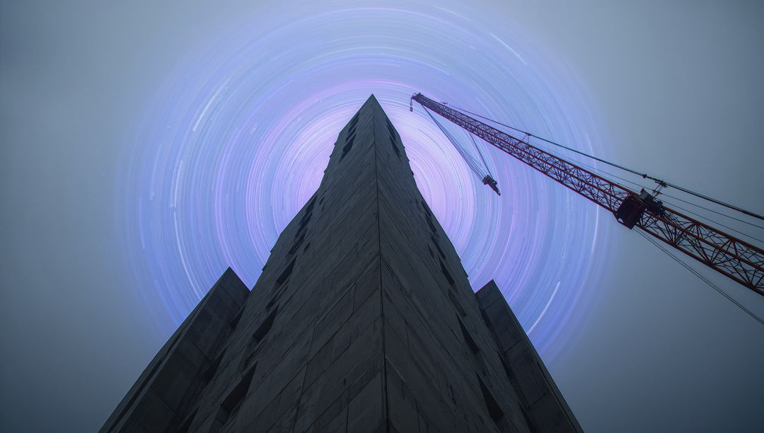 Soaring concrete tower apex with red crane and circular star trails at night
