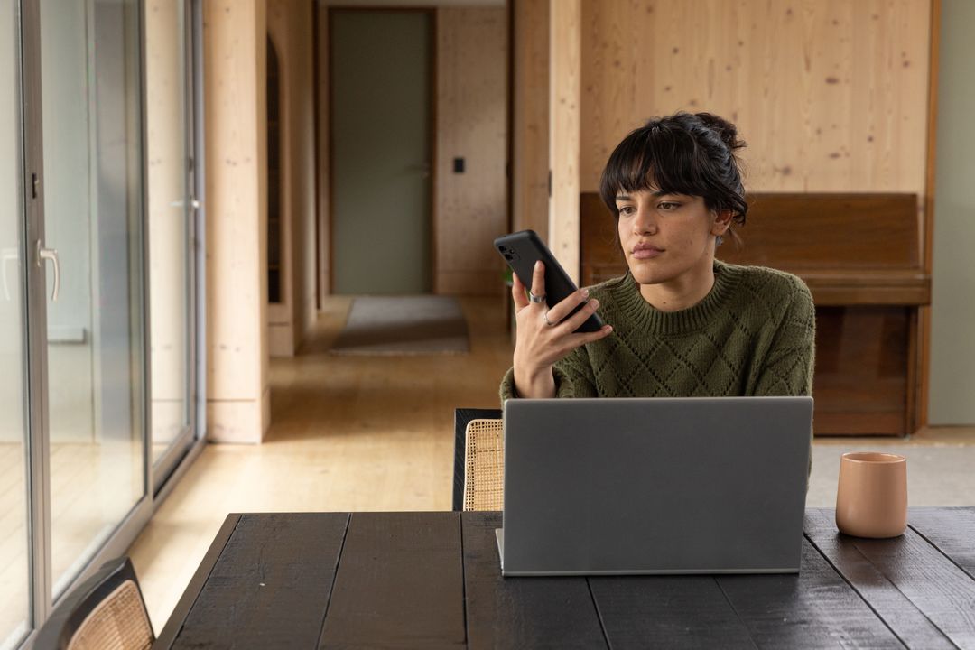 Woman Engaging with Smartphone While Working on Laptop at Cozy Home