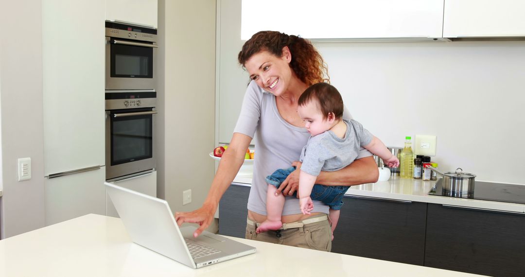 Multitasking mother working on laptop while holding baby in modern kitchen - Free Images, Stock Photos and Pictures on Pikwizard.com