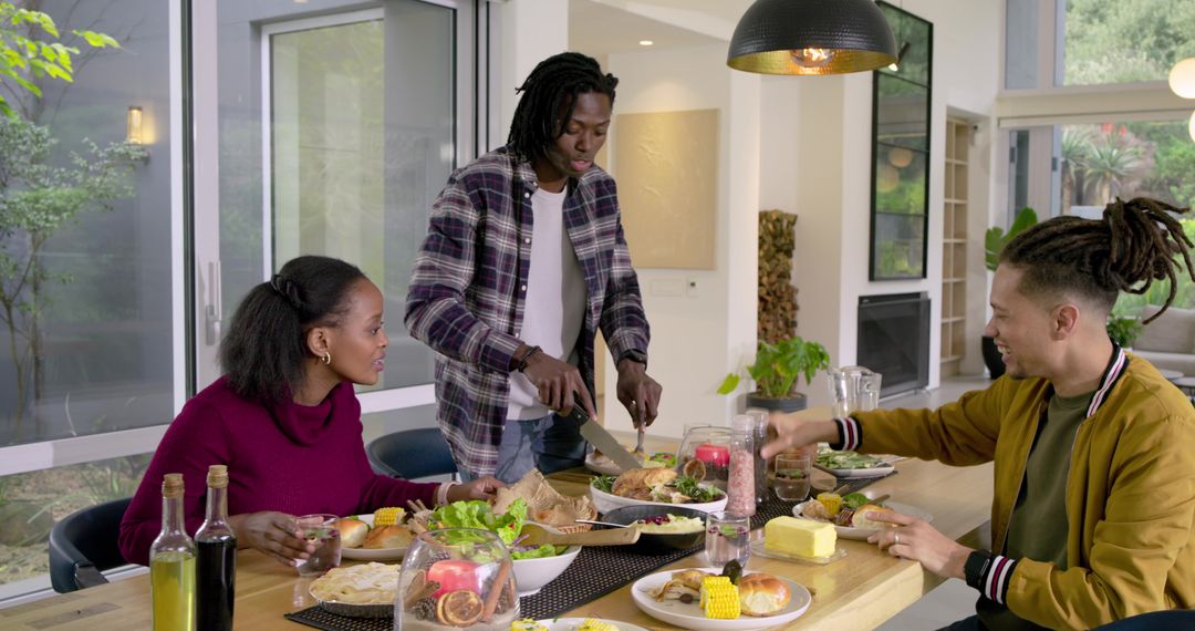 Diverse friends sharing family-style dinner with man carving chicken at modern dining table