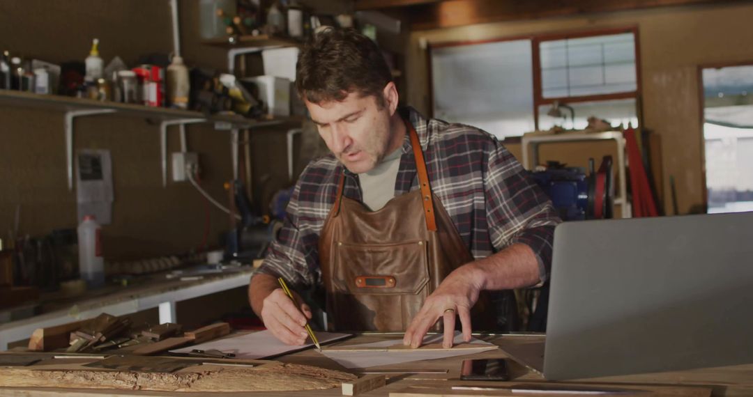 Mature craftsman marking woodworking plans at rustic workbench with laptop and tools