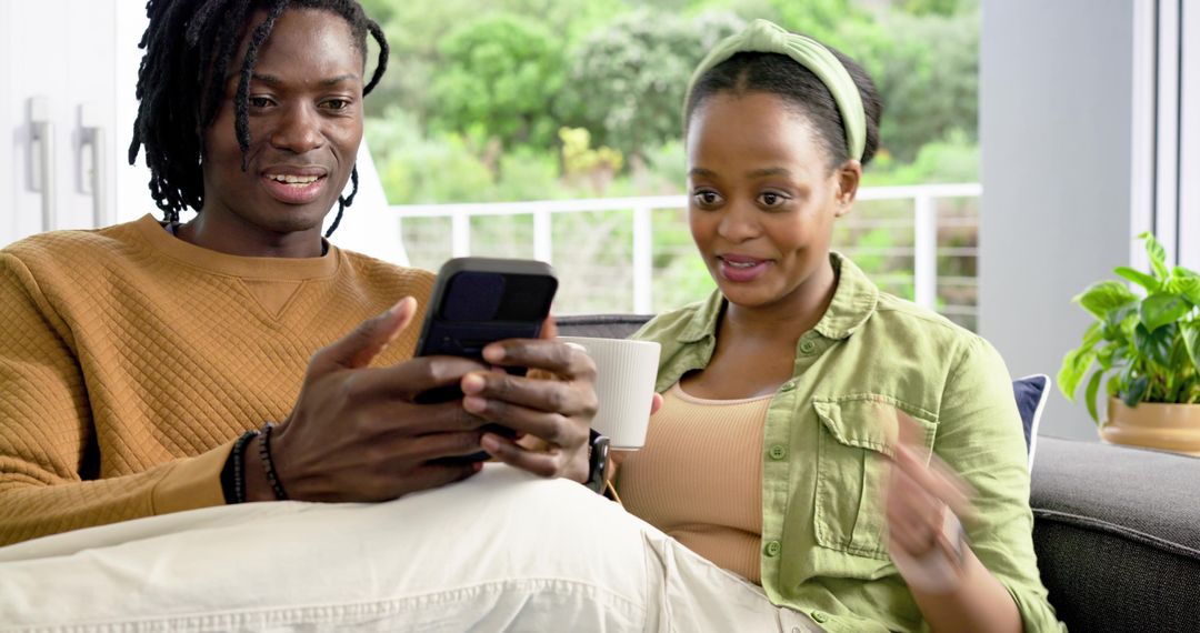African American Couple Sharing Flip Phone and Coffee on Sofa, Laughing and Relaxing