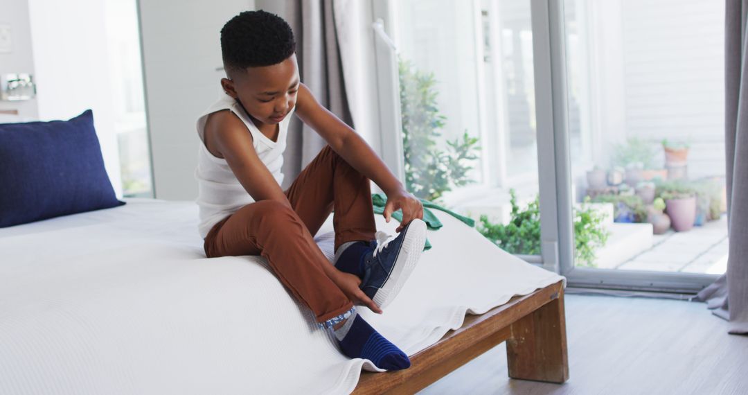 Young Boy Sitting on Bed Tying Sneakers by Windows - Free Images, Stock Photos and Pictures on Pikwizard.com