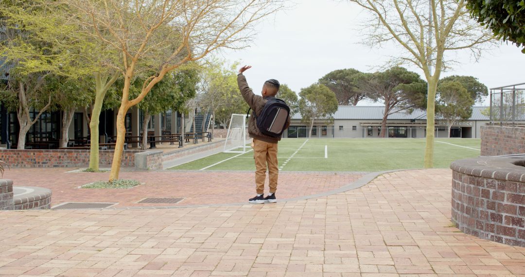 Child Waving in Schoolyard with Backpack and Sports Field