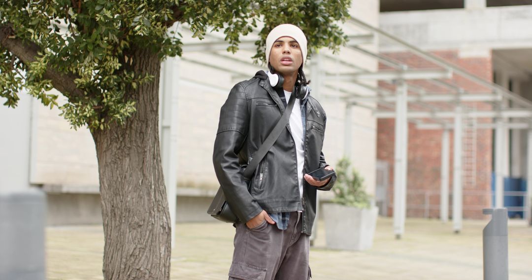Urban student standing with smartphone and headphones in overcast campus courtyard, wearing beanie