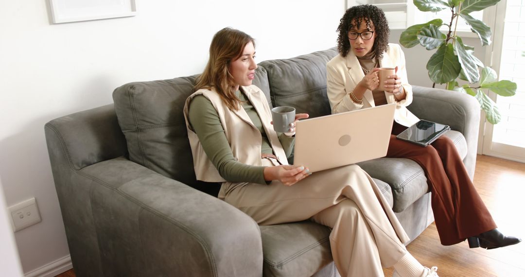 Two Female Colleagues Collaborating on Laptop and Tablet on Sofa with Coffee at Home