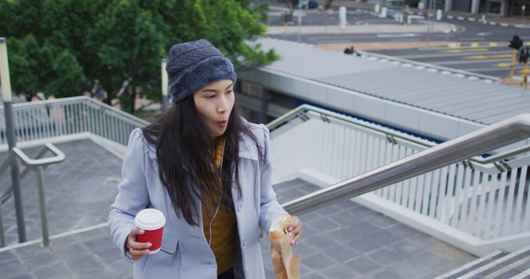 Asian woman walking holding takeaway coffee and eating sandwich - Free Images, Stock Photos and Pictures on Pikwizard.com