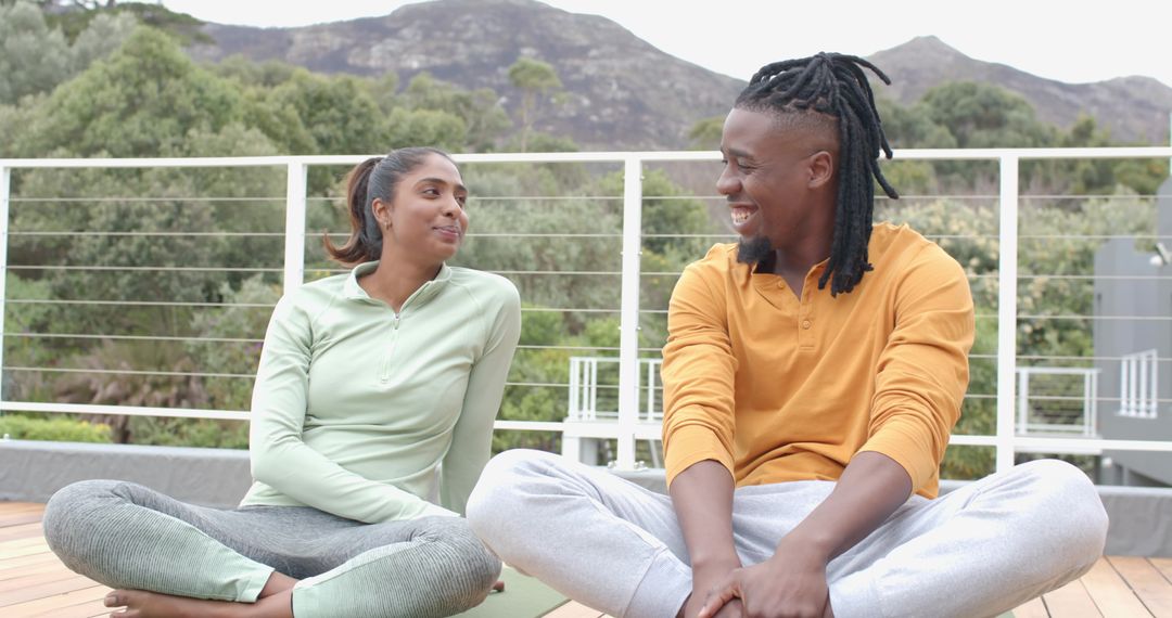 Diverse couple practicing yoga and chatting on outdoor deck with mountain and greenery backdrop