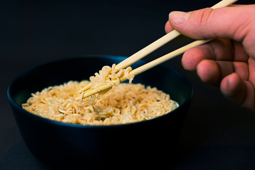 Hand Using Chopsticks Lifting Instant Ramen Noodles from Black Bowl Close-Up