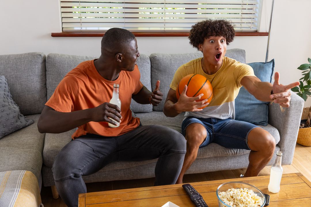 Multiethnic friends cheering on sofa holding basketball and bottles while grabbing popcorn