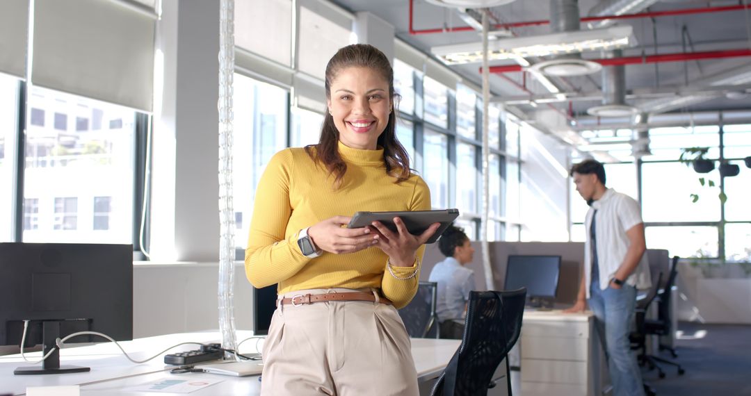 Female professional holding tablet smiling in bright modern open office with colleagues collaboratin