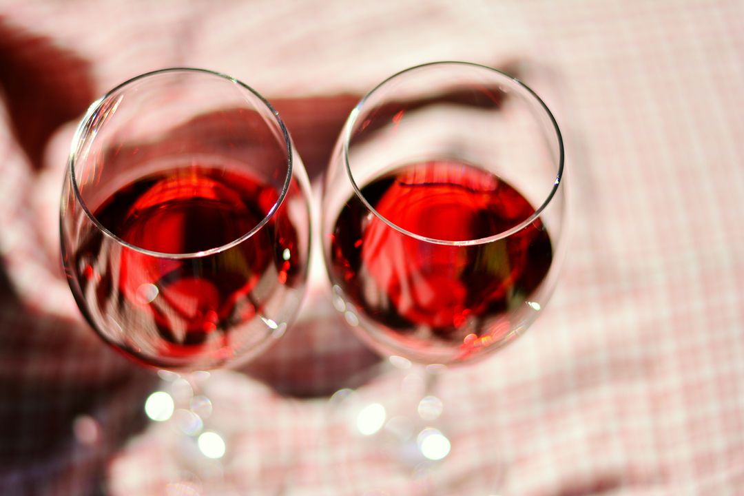 Two glasses red wine resting on pink gingham tablecloth under warm sunlight top view
