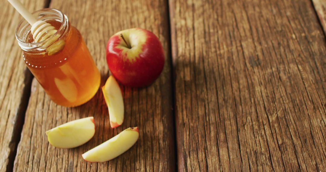 Rustic honey jar with wooden dipper and red apple slices on weathered wood surface