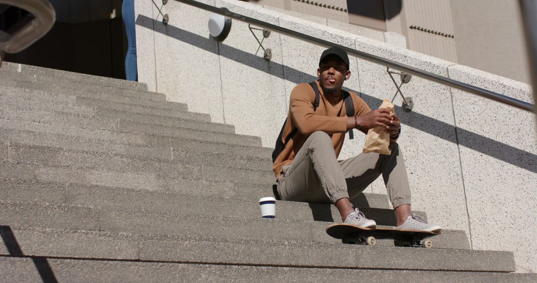 African American student sitting on steps eating lunch with skateboard and backpack