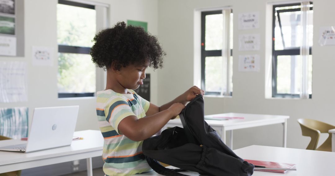 Elementary student packing backpack at classroom table with laptop and notebooks