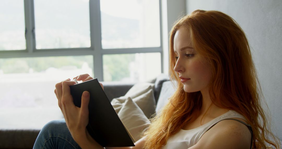 Red-haired woman reading book by window in bright living room - Free Images, Stock Photos and Pictures on Pikwizard.com