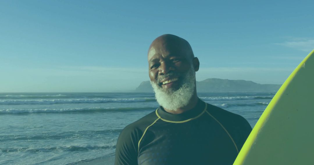 Senior surfer smiling while holding bright surfboard on sunlit ocean shore at sunrise