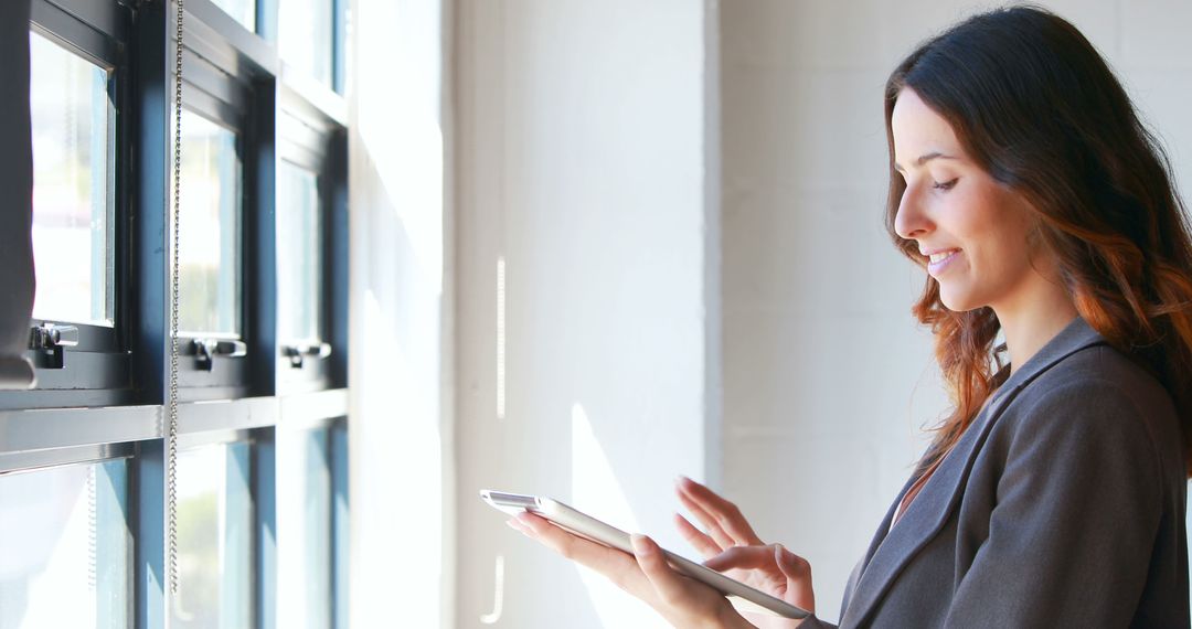 Businesswoman Using Tablet in Office by Window with Smile - Free Images, Stock Photos and Pictures on Pikwizard.com