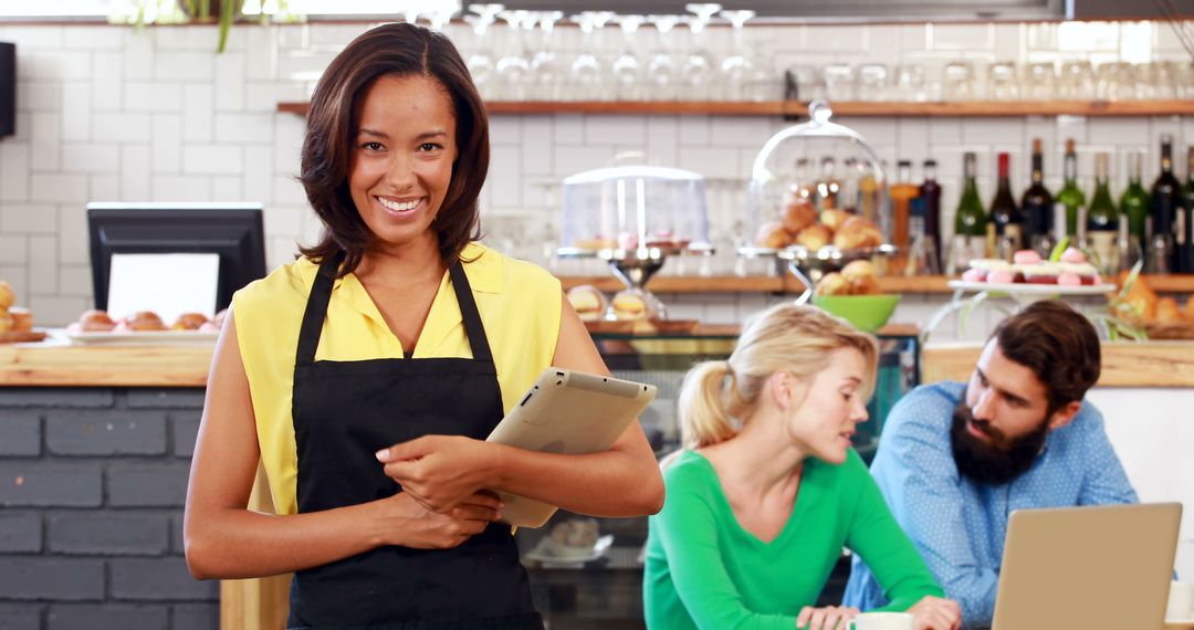 Smiling Café Worker Holding Digital Tablet with Customers in Background - Free Images, Stock Photos and Pictures on Pikwizard.com