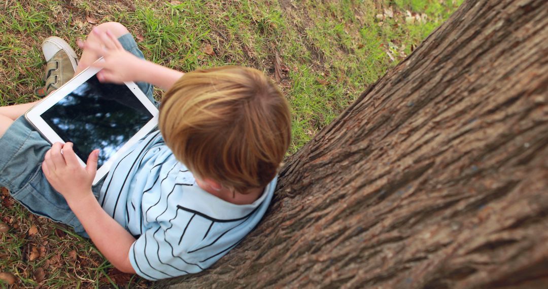 Young Boy Using Tablet While Leaning Against Tree in Park - Free Images, Stock Photos and Pictures on Pikwizard.com