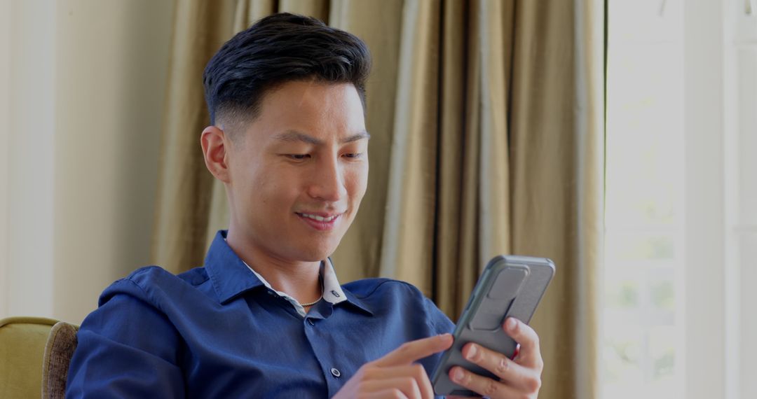 Contemplative Asian Man Relaxing with Smartphone by Window