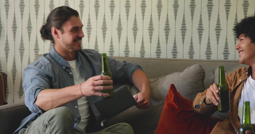 Two friends relaxing on couch sharing beer and laughs with tablet for casual home hangout