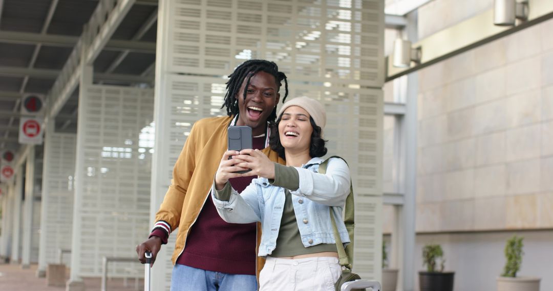 Multicultural couple taking selfie in modern transit concourse holding smartphone and rolling luggag