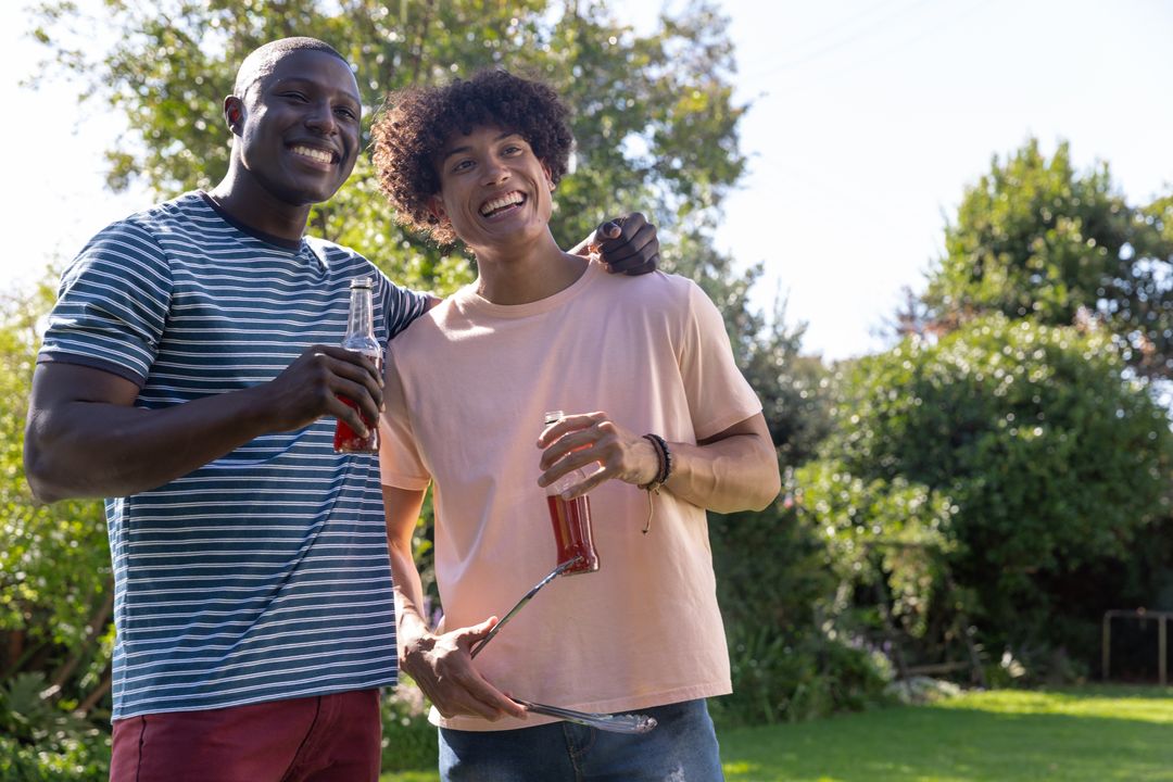 Smiling diverse friends enjoying sunny backyard barbecue while holding drinks and grilling tongs