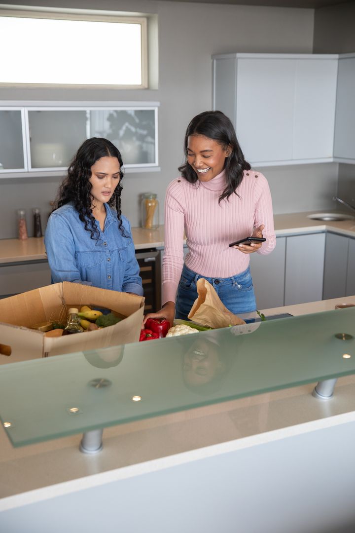 Diverse Friends Unpacking Grocery Box Using Smartphone Kitchen Counter