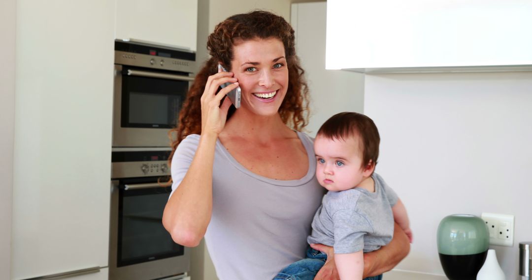 Happy Mother Holding Baby While Talking on Phone in Kitchen - Free Images, Stock Photos and Pictures on Pikwizard.com