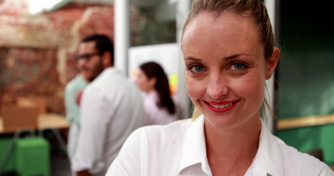 Smiling young woman in office setting with colleagues in background - Free Images, Stock Photos and Pictures on Pikwizard.com