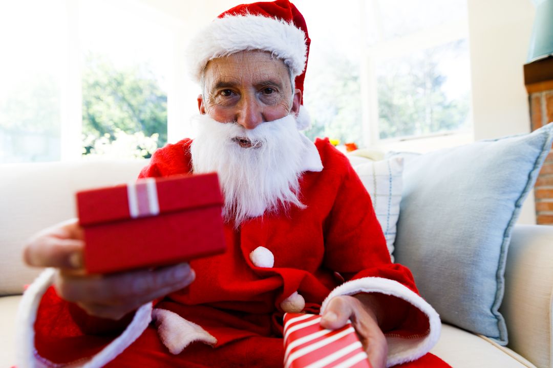 Elderly man in Santa costume presents gift while seated on couch. - Free Images, Stock Photos and Pictures on Pikwizard.com