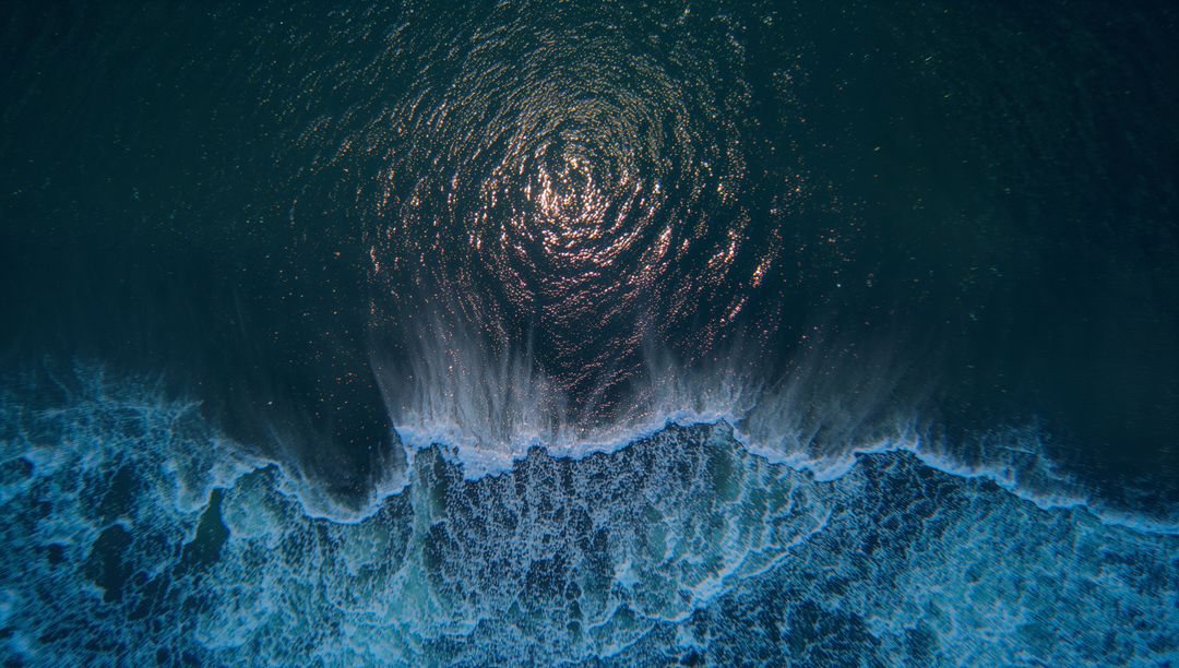 Aerial drone view breaking surf forming white foam and sun-glint ripples across blue ocean