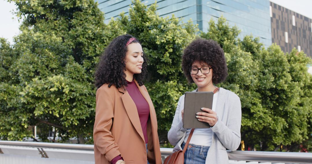 Diverse women smiling while sharing tablet and collaborating on urban bridge with greenery